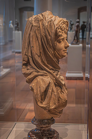 A detailed sculpture of a draped figure on display, encapsulated in a glass case within an National art gallery, surrounded by other sculptures in the background.