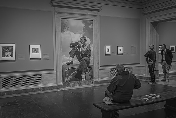 An art gallery visitor seated in contemplation, others standing and discussing the photography, with Dorothea Lange’s image as the central piece, at the Lange exhibition.