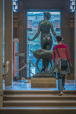 A visitor at the National Gallery admiring a classical bronze sculpture by a grand entrance, with daylight accentuating the form of the artwork