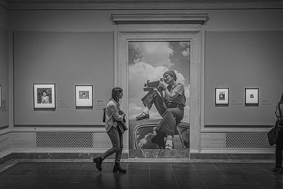 An exhibition hall with a visitor viewing Dorothea Lange's photographic works, including a large central photo of Lange with her camera, and surrounding framed black-and-white portraits.