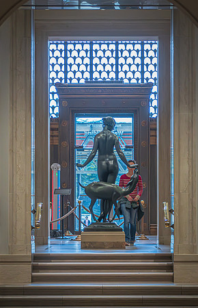 A sculpture positioned at the entrance of the National Gallery, illuminated by natural light from a decorative window, greets visitors.