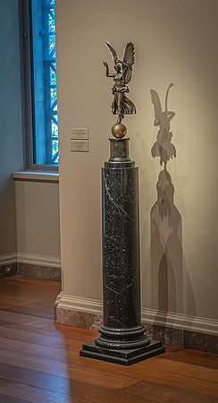 Bronze winged sculpture on a marble pedestal with its shadow cast on a wall in the National Gallery, near a window with a blue grid pattern.