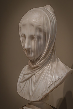 A sculpture of a veiled person intricately carved in white marble, displaying the texture of fabric and the subtlety of the figure's face beneath.