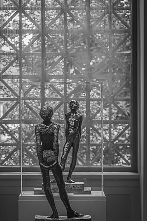 Two slender bronze sculptures displayed before a geometric lattice window, basking in the diffused light within an art gallery setting.