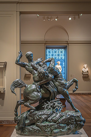 A dynamic bronze sculpture depicting mythological figures on a rearing horse, set against the backdrop of an art gallery with soft natural light filtering through a window.