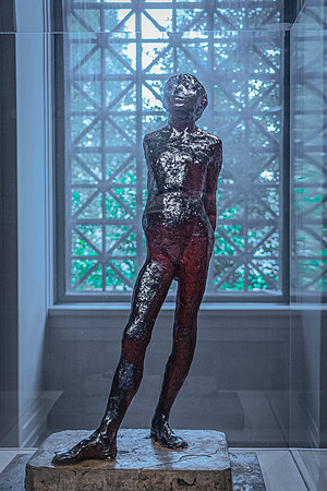 A bronze sculpture on display at the National Gallery, highlighted by natural light casting dramatic shadows against a geometric-patterned window backdrop.
