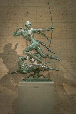 A bronze statue of Artemis, Greek goddess of the hunt, poised with a bow, in the National Gallery, against a textured wall backdrop.